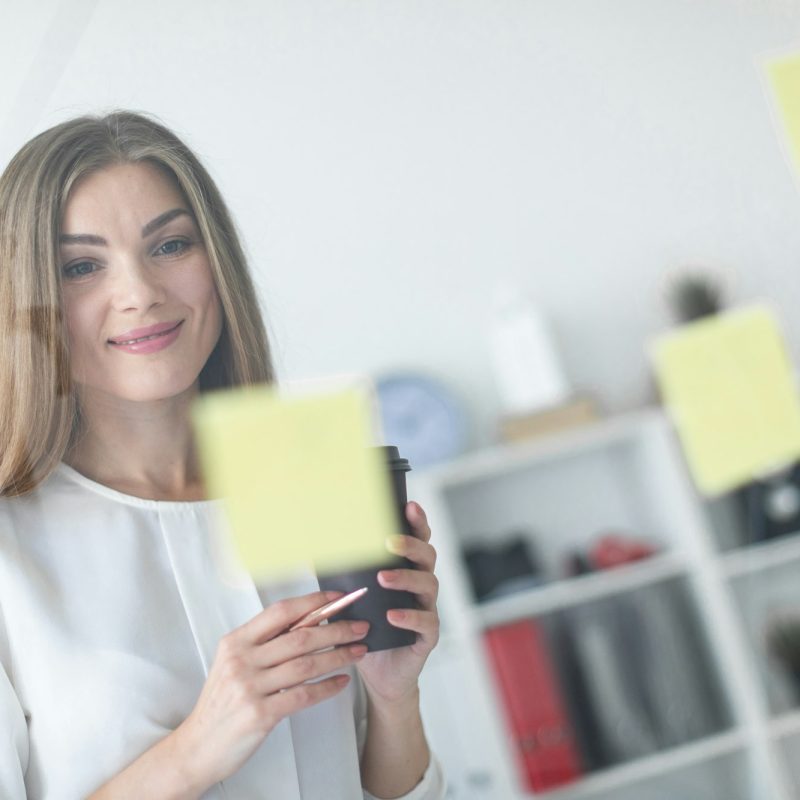 A young girl stands near a transparent board with stickers and holds a glass with coffee and a pen.